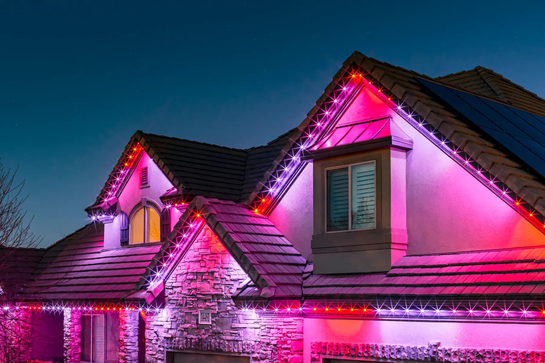 Wide shot of a home outlined in red, green and pink permanent roofline lights at night